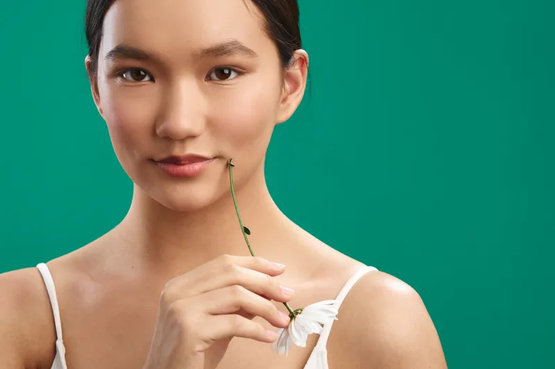 Asian woman in a white top holds a flower near her lips against a teal backdrop, creating a calm, elegant mood and a soft, natural portrait that emphasizes subtle beauty and poise.