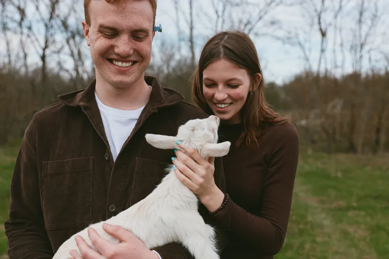 Couple outdoor lifestyle scene with a goat kid, animal affection, happy smiles, candid moment as they cradle the young goat in a sunny park setting, surrounded by trees and open grass.
