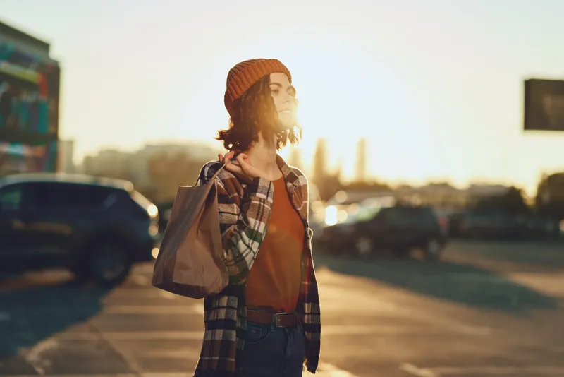 Woman shopping in urban street at golden hour, candid lifestyle portrait with beanie and bag, warm backlight and golden hour glow, authenticity, mindful living, emotional storytelling.