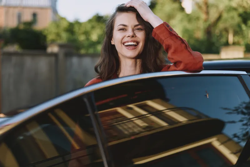 Smiling young woman with long dark hair leans from a car window, enjoying a sunny, carefree moment as the vehicle reflects surrounding buildings