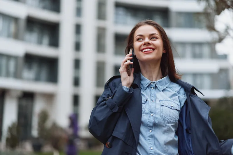 Young brunette woman talks on her cellphone outdoors, wearing a light denim shirt under a dark jacket, smiling as the urban background frames her confident pose.