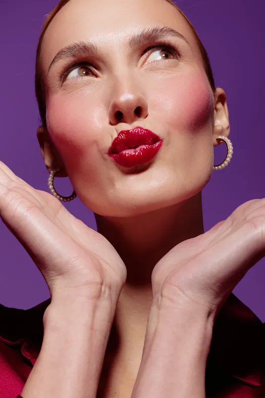 Close-up of a woman with red lipstick making a playful kiss face, hands framing her face against a purple background. Concept of beauty, makeup, and fun expression.