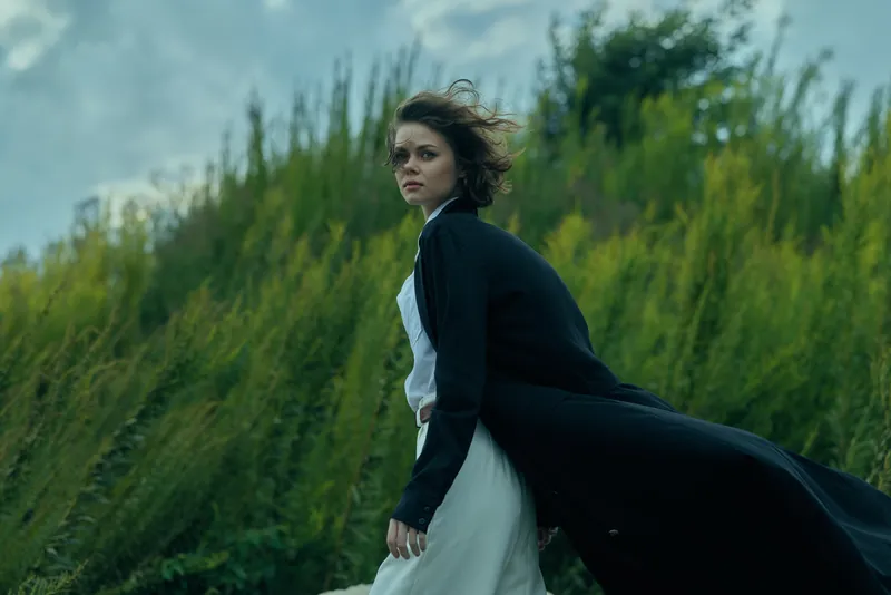 Woman in a flowing dress and dark coat stands amid tall grasses, wind sculpting her silhouette in a dramatic outdoor moment, cinematic and atmospheric