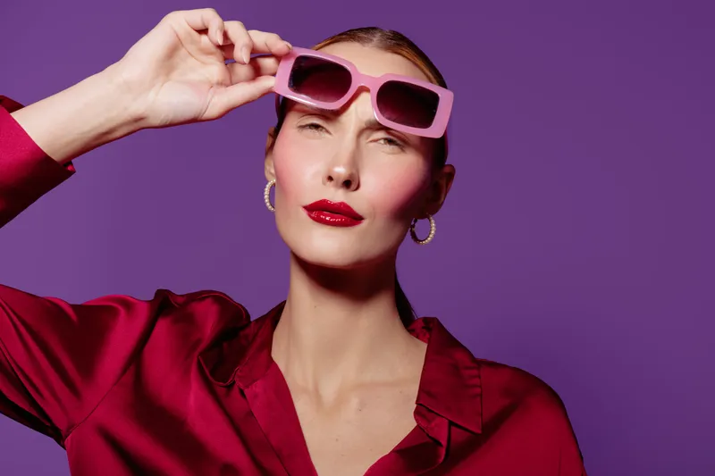 Stylish woman in red satin blouse adjusts pink sunglasses on purple background. Fashion portrait with glossy red lips and hoop earrings. Concept of glamour, confidence, and modern style.