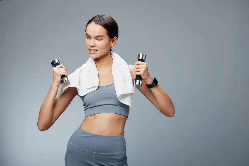 Focused woman in athletic wear lifts dumbbells with a towel over shoulders, mid workout in a clean studio, showcasing strength, balance and determination during a gym session.
