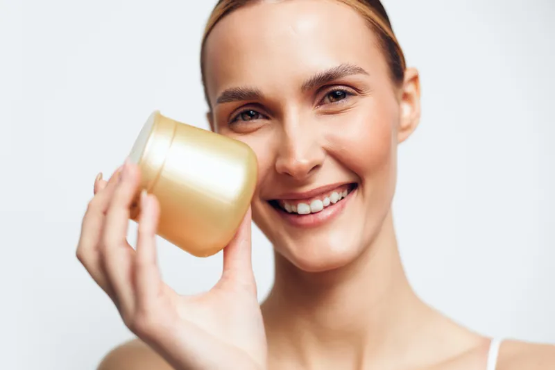 A smiling young woman holds a golden cosmetic jar close to her face against a white background. The image represents skincare, beauty, wellness, and personal care concepts.