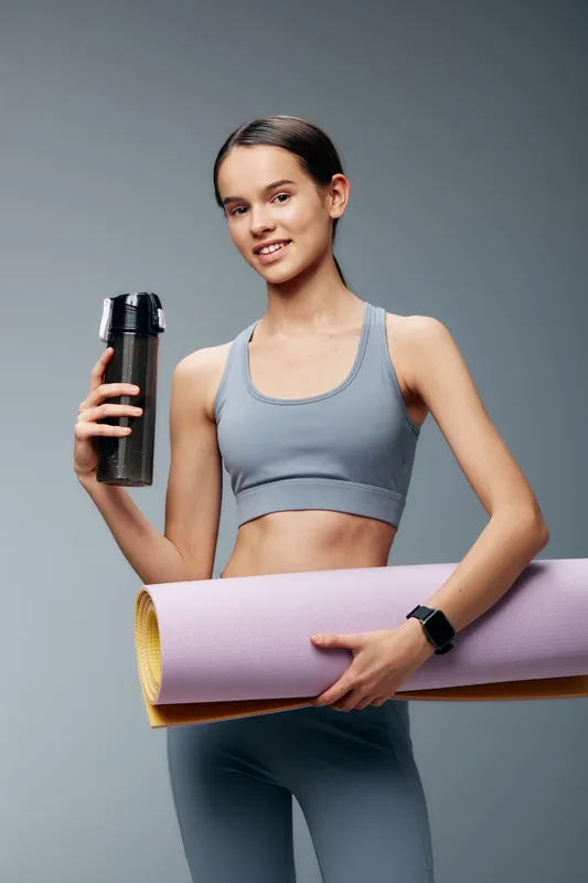 Female fitness enthusiast in a light blue sports set, holding a water bottle and rolled yoga mat, standing in a studio ready to start a workout and healthy routine.