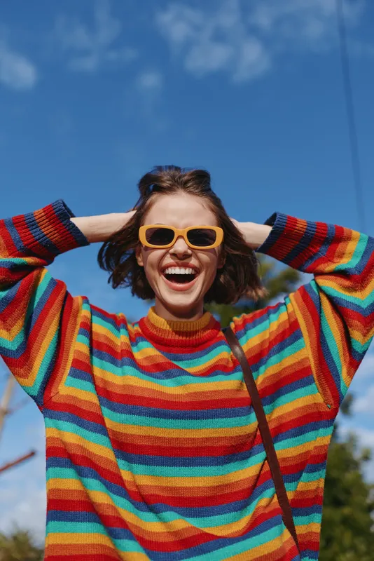Woman smiling in colorful striped sweater and yellow sunglasses, hands behind head outdoors under blue sky. Joyful portrait of young adult in casual fashion on a sunny day.