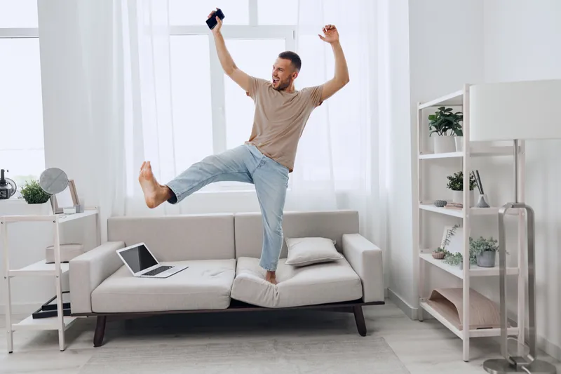 Happy man celebrating at home, dancing joyfully with his smartphone, surrounded by a bright, modern living space filled with plants, showcasing a vibrant lifestyle.