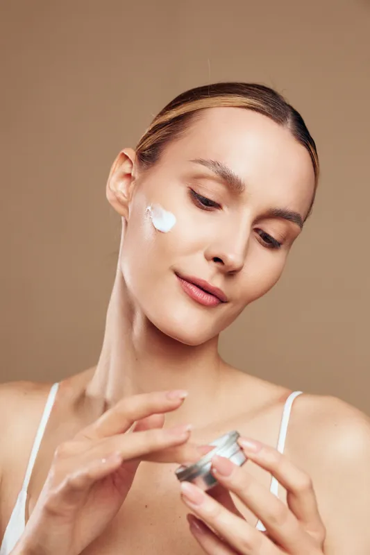 Young woman applying face cream, skincare routine, natural beauty, soft lighting, clear skin, calm expression, beige background, close up portrait, self care moment