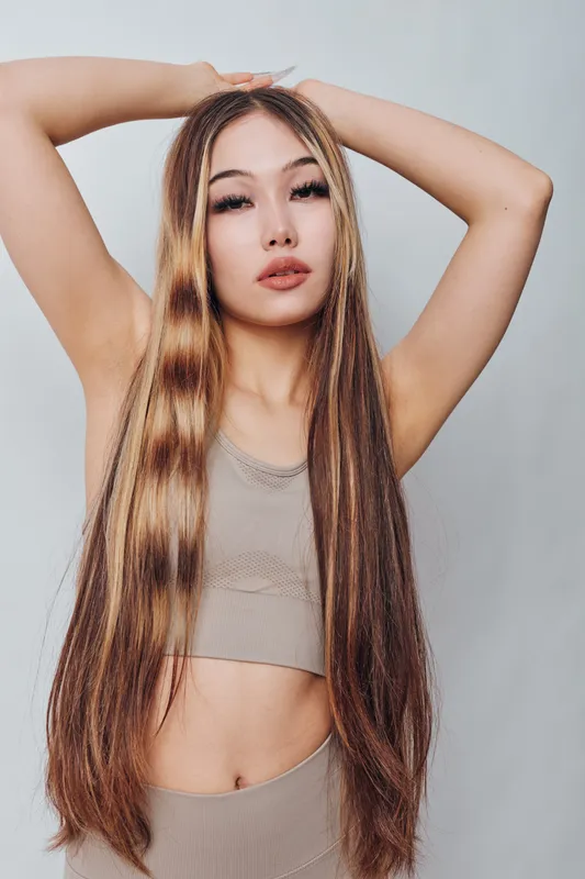 Young woman posing with long straight hair and arms raised wearing crop top and leggings. Beauty, fashion, and hairstyle with natural makeup on neutral gray background.