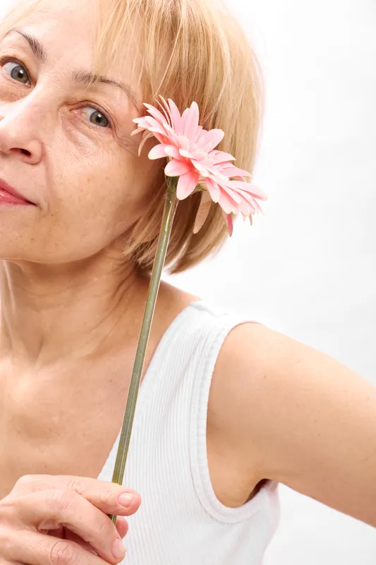 Smiling woman holds a pink flower near her face, closeup on white studio background, soft lighting, concept of skincare, beauty, wellness, selfcare, calm
