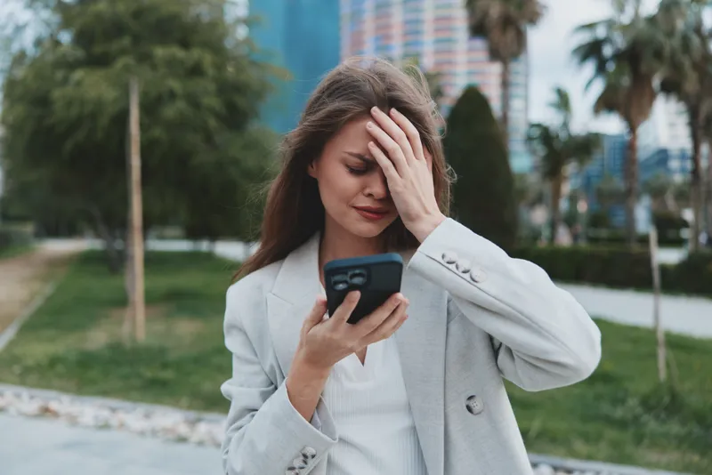 Woman stressed by financial crisis reading stock market fall news on smartphone in urban outdoor setting with green palms and buildings