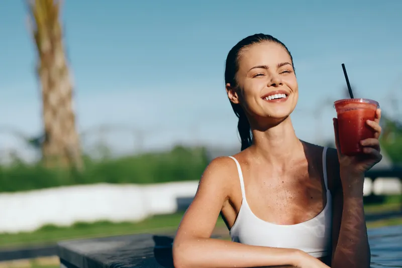 Cheerful woman outdoors, smiling with sunlit glow, casual tank top, refreshing drink in hand, relaxed posture, vibrant colors, palm trees in background, summer lifestyle moment