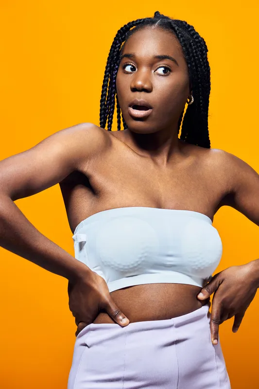 Surprised young Black woman in a white strapless top and pastel trousers, looking curiously with hands on hips against a vibrant yellow background