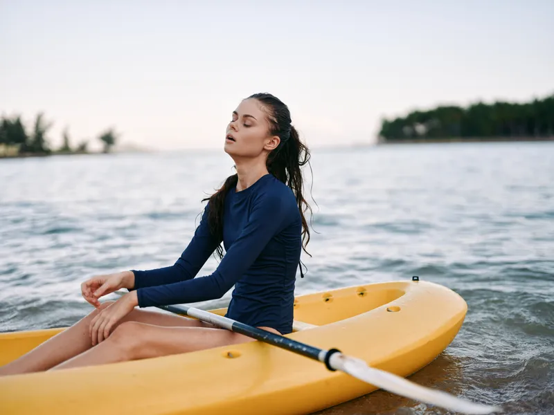 A calm figure in a navy long sleeve top sits in a yellow kayak on gentle water, eyes closed, enjoying peaceful isolation and reflective moment by the shore.