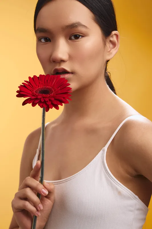 Asian woman holds a bold red flower close to her face, standing against a warm yellow background. The composition exudes elegance, calm charm, and a minimalist, artistic mood.