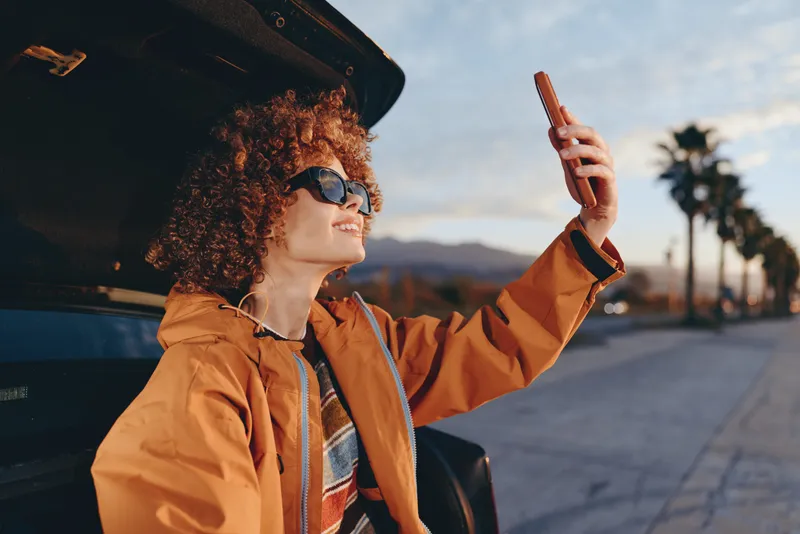 Woman with curly hair wearing sunglasses and an orange jacket sits by a car door outdoors, raising a smartphone for a selfie. Travel lifestyle, warm sunset light, distant mountains.