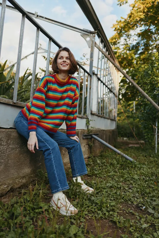 Woman in colorful striped sweater and jeans sitting outdoors by a metal railing, smiling and looking up, casual portrait of a relaxed young adult in a green garden setting.