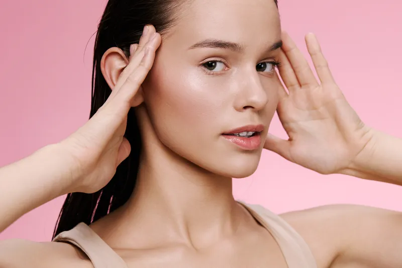 A young woman with long dark hair gently touching her face, showcasing a natural makeup look against a soft pink background, radiating beauty and confidence