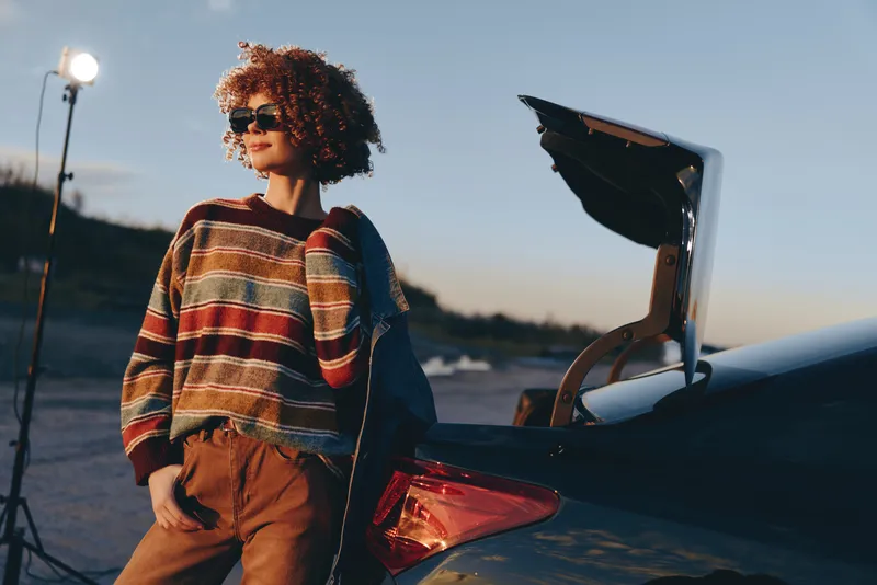 Woman lifestyle portrait with curly hair wearing sunglasses beside a car trunk, laptop open with a graduation cap, outdoors at sunset, casual travel mood, soft evening light, natural breeze