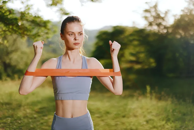Fit young woman exercising outdoors with resistance band during summer training session in nature, showcasing strength and determination while enjoying fitness