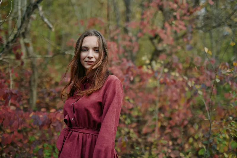 Portrait of a young woman in a burgundy coat amid autumn foliage, natural pose and soft smile conveying authenticity and credibility, captured with candid lighting and true-to-life tones.