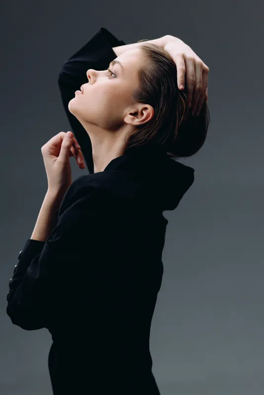 Elegant woman posing with hand in hair against dark gray background, showcasing stylish black clothing and calm expression.