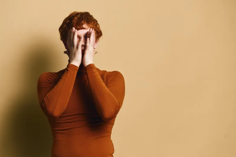 Portrait of young woman with curly red hair covering her face with hands against beige wall. Casual style, isolated, expression and self concealment concept with warm colors and minimal background.