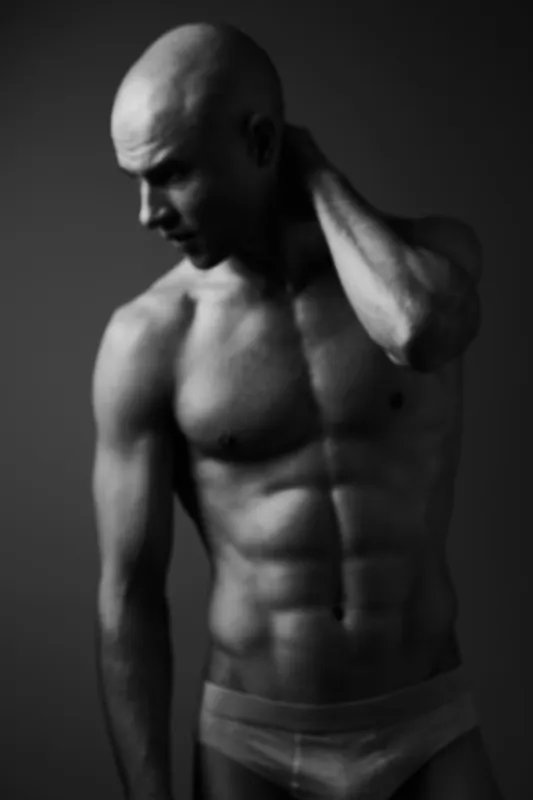 Black and white image of a muscular man striking a contemplative pose, showcasing strength and emotion. The simple background enhances the focus on his physique.