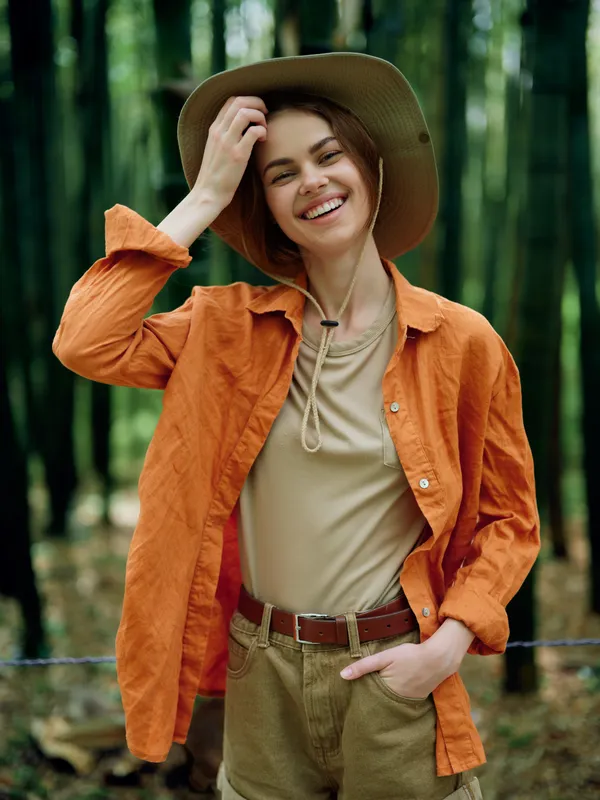 Woman with smile and hat in forest, orange jacket and casual outfit for portrait, happy young traveler posing relaxed with hand in pocket, joyful outdoor lifestyle and nature escape.