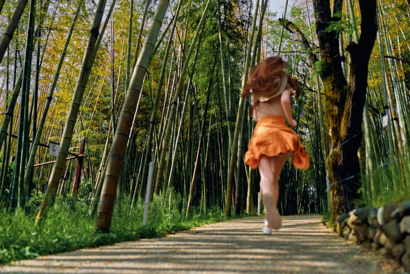 Woman running on a sunlit bamboo forest path in an orange skirt, a back view capturing motion and joyful movement along a leafy trail surrounded by tall green trees.
