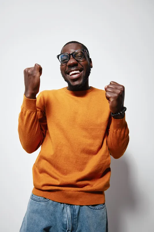 African American man with dark skin wearing glasses and orange sweater celebrating success with clenched fists and joyful smile on white background in studio.