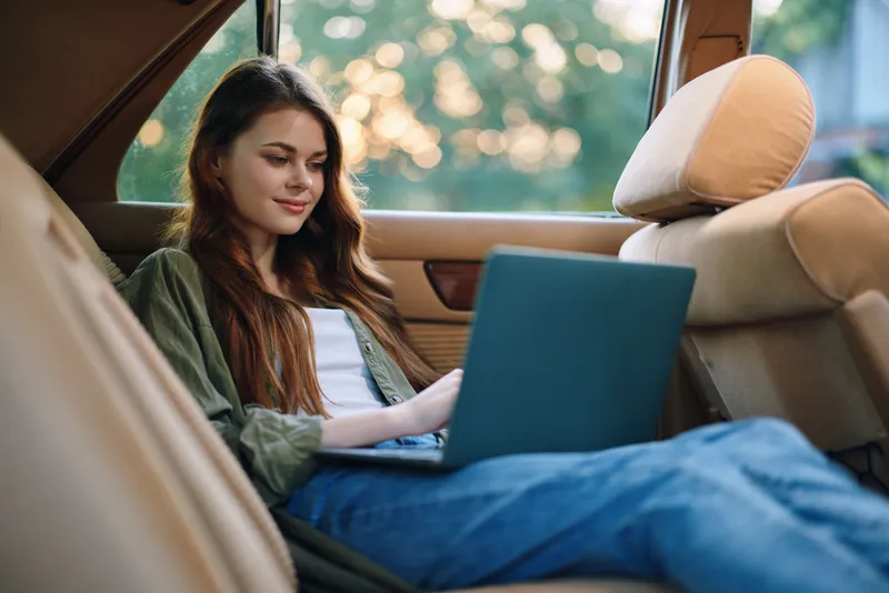 Young woman working on a laptop in a car, enjoying a casual and relaxed atmosphere during a warm evening.