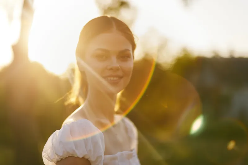 Smiling young woman in a white dress during golden hour, vibrant sunlight creating a warm atmosphere and joyful emotion in a natural setting