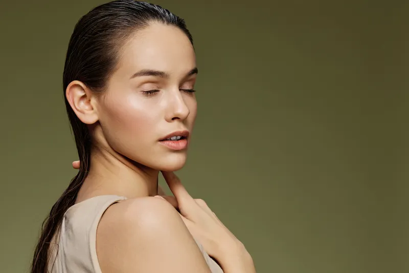 Close up portrait of a serene female model with wet hair, wearing a beige top, set against a calming green background conveying tranquility and natural beauty
