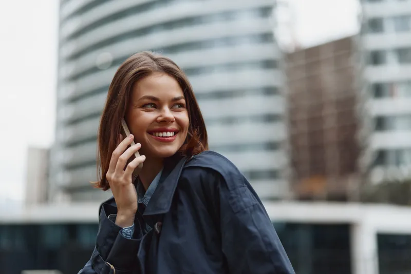 A smiling young adult woman with brunette hair talks on a phone in a busy urban street, wearing a navy denim jacket, with modern glass buildings in the background