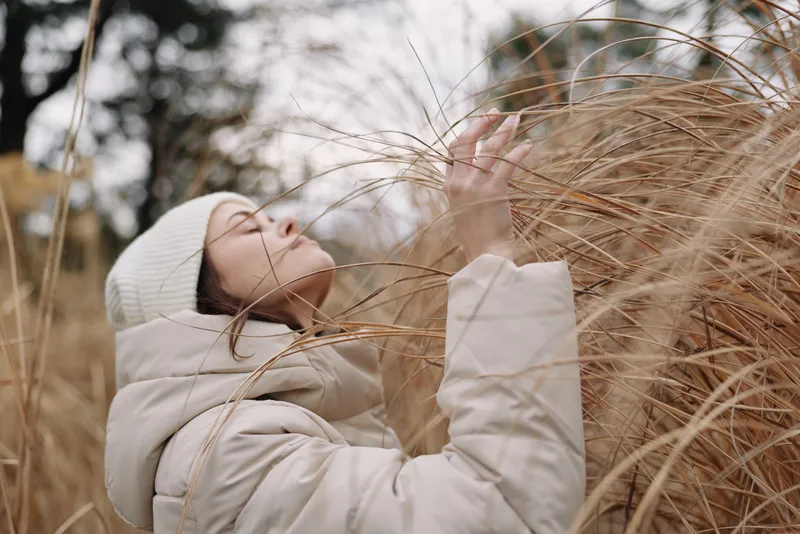 Winter woman in a warm coat outdoors touching tall grass, a calm moment in nature with gentle wind and soft light.