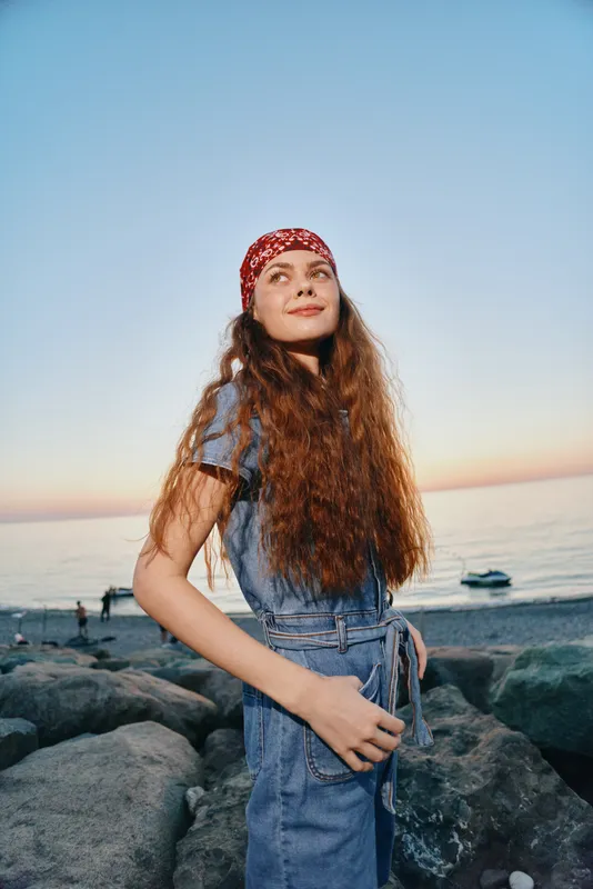 Lifestyle woman in modern boho-western denim street style standing on rocky beach at sunset, film color tones enhance warm natural mood and casual fashion look