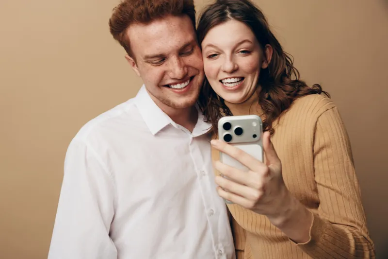 Happy young couple smiling and taking a selfie with smartphone on beige background. Casual clothing, joyful mood, close relationship, modern technology, and social connection concept.