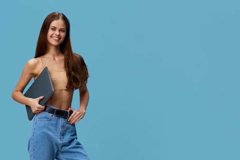 smiling woman holding laptop wearing casual beige top and blue jeans with long brown hair standing against blue background