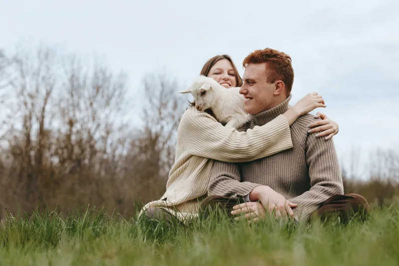 A couple sits on the grass in a meadow with a friendly goat kid, sharing affection outdoors, capturing a warm lifestyle moment and natural bond.