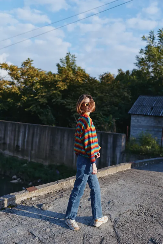 Young woman in striped sweater and sunglasses walks on a concrete rooftop edge, smiling and looking back; casual jeans and sneakers, short bob haircut, sunny urban travel lifestyle portrait.
