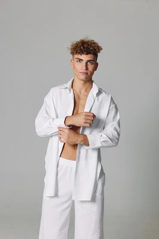 Young man with curly hair wearing white open shirt and pants posing against gray background, casual style and confident expression in studio portrait.