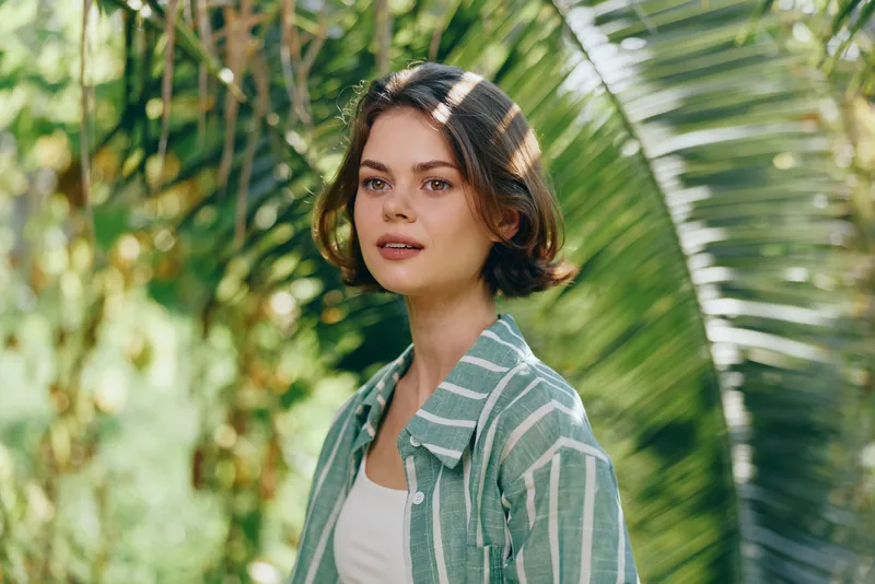 Portrait of a young woman outdoors among lush greenery, wearing a striped green shirt, soft natural light and calm expression.