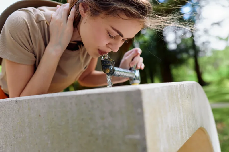 girl drinking water from a stone fountain in a park, outdoor closeup of a young woman sipping from the spout, summertime hydration and candid portrait showing face, hands and flowing stream