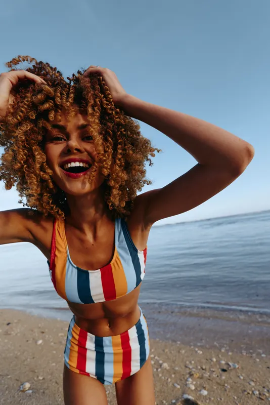 Joyful woman in striped swimsuit smiles, windblown hair by the beach and calm ocean.