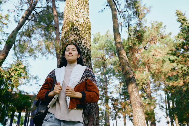 Woman outdoors through a forest scene, wearing a knitted jacket and backpack, strolling under tall trees as autumn light creates calm mood and lifestyle in nature