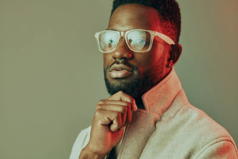 Stylish Black man in glasses, wearing a fashionable coat, with a thoughtful expression and a minimalistic background, ideal for fashion and lifestyle themes