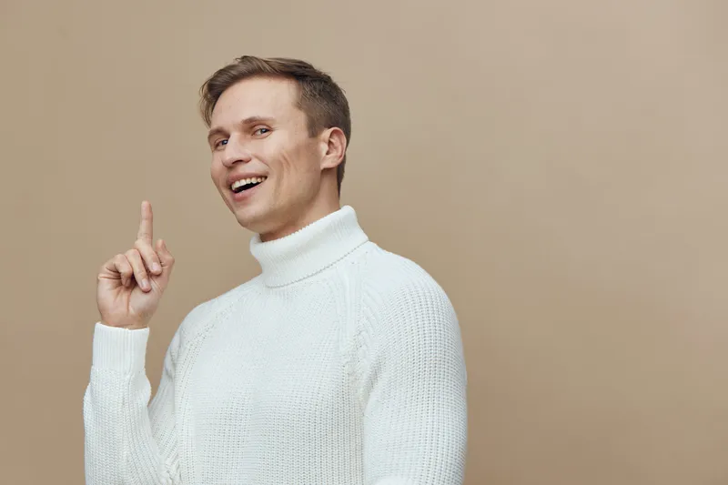 cheerful smiling caucasian man wear white knitted sweater pointing finger up isolated beige background indoor portrait digital detox concept